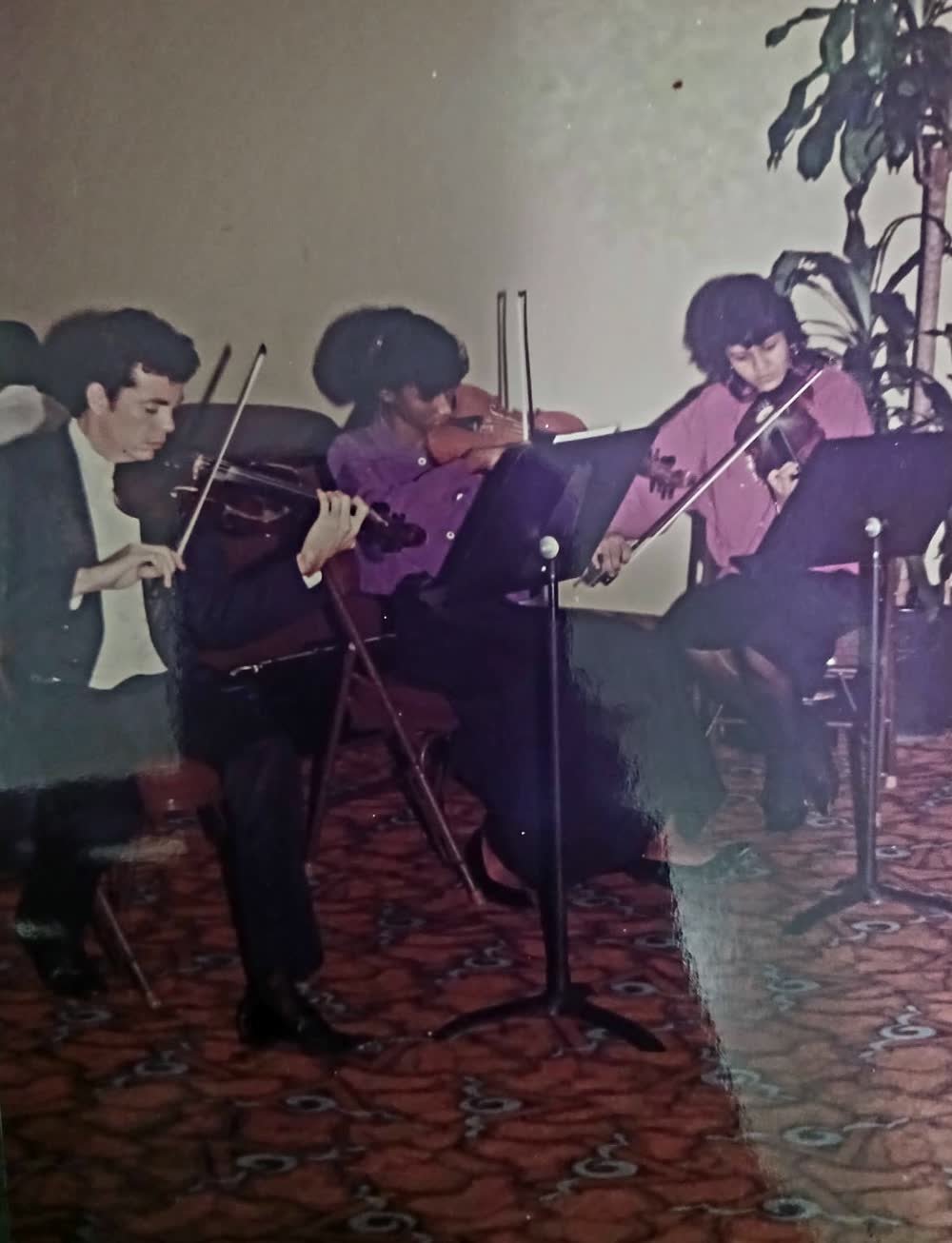 Melanie joven, con blusa rosada, tocando el violín en una orquesta de cámara