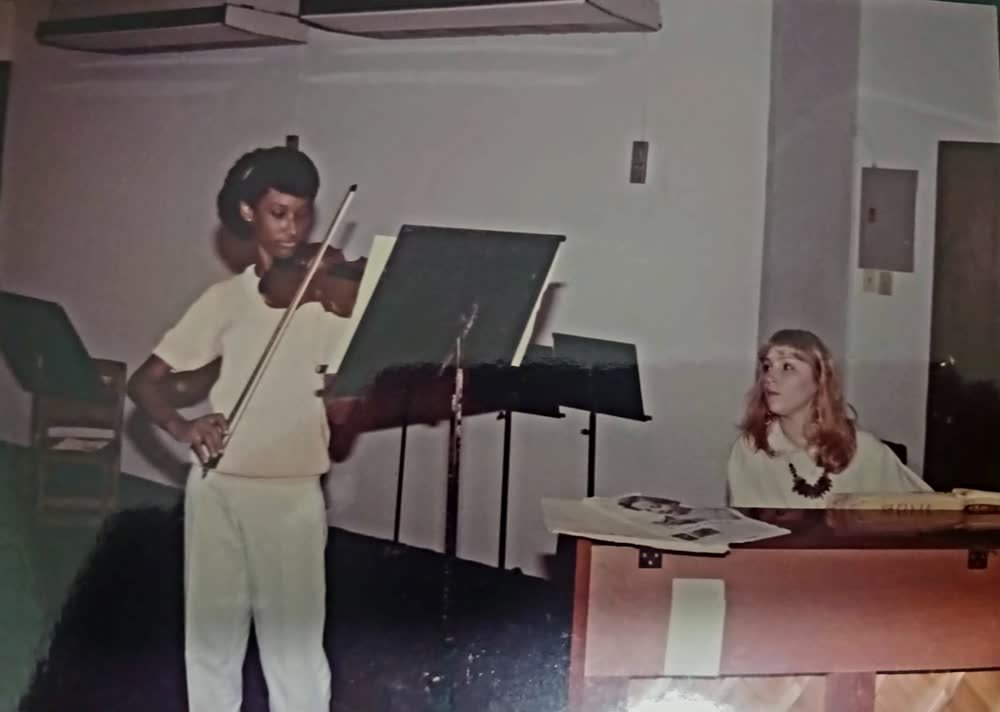 Melanie adolescente tocando el violín junto a su profesora al piano